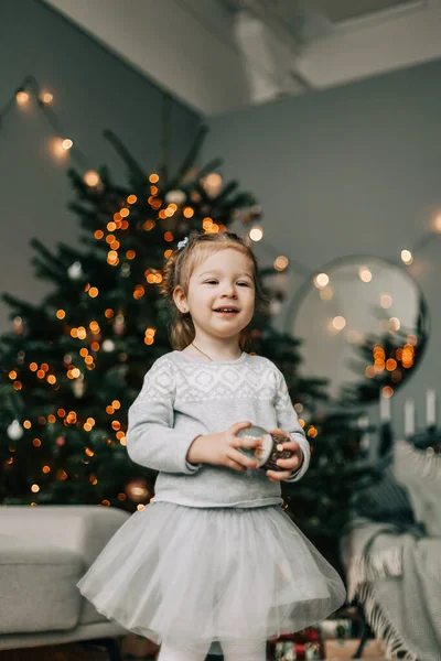 A beautiful little girl in a festive dress is standing at the Christmas tree. the concept of Christmas and New Year — Stock Photo, Image