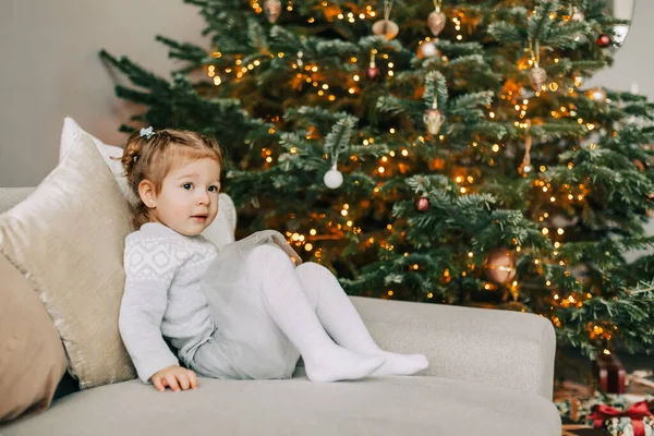 A pretty girl in a hat is sitting on the sofa near a Christmas tree, dressed up for the holiday, the child is smiling and rejoicing at the gift — Stock Photo, Image