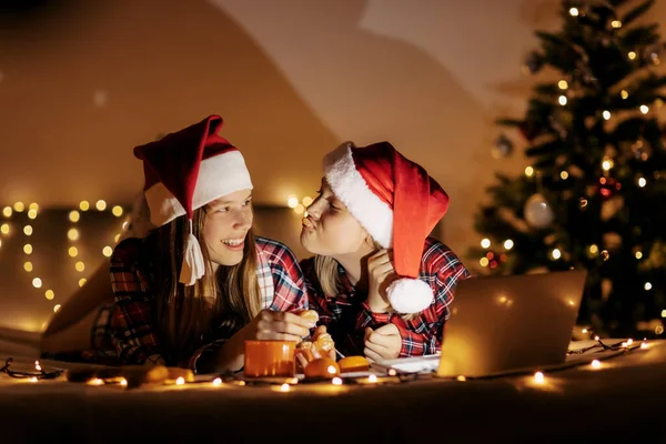 Due ragazze adolescenti in camicia a quadri sdraiate sul letto e con un computer portatile, un albero di Natale illuminato sullo sfondo. Atmosfera natalizia, periodo natalizio, tempo di magia — Foto Stock