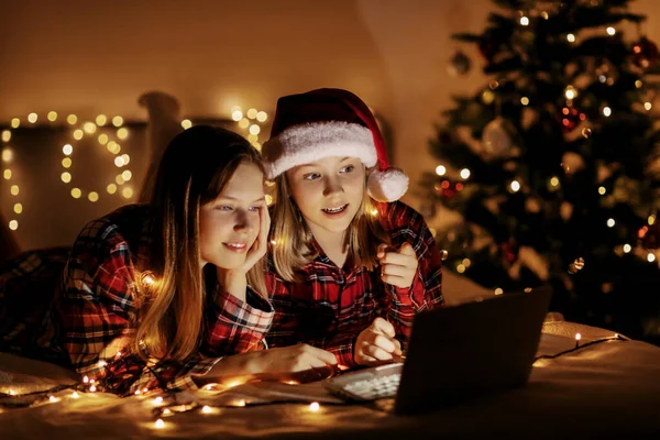 Dua saudara perempuan yang menawan berbaring di tempat tidur dengan laptop pada malam Natal atau Tahun Baru dan mengucapkan selamat kepada teman-teman dan kerabat mereka secara online. Salam Natal online, pesta virtual untuk Natal Stok Foto Bebas Royalti