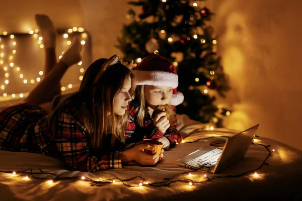 Dua gadis remaja dengan kemeja kotak-kotak berbaring di tempat tidur dan menggunakan laptop, pohon Natal terang di latar belakang. Natal suasana, waktu Natal, waktu sihir Stok Lukisan  