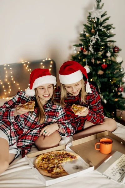 Due ragazze affascinanti in cappelli di Babbo Natale e camicie a quadri sono seduti sul letto e si godono la pizza ordinata, guardando un film durante le vacanze di Natale. — Foto Stock