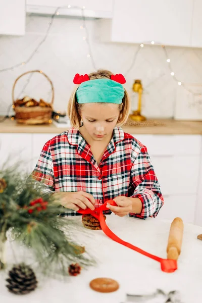 Una chica bonita decora galletas con una cinta roja brillante, prepara un dulce para los huéspedes en Navidad o Nochevieja — Foto de Stock