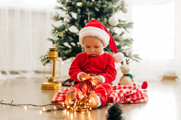 Un niño alegre en un disfraz de Año Nuevo se sienta en el suelo cerca del árbol de Navidad y juega con una guirnalda de Navidad — Foto de Stock