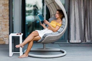 Genç bir kadın terasta asılı bir sandalyede oturmuş dergi okuyor.