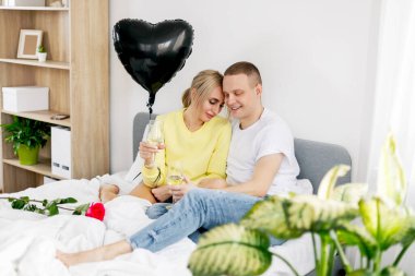 Genç bir adam ve bir kız yatak odasında bir kadeh şarap, bir balon ve çiçeklerle sevgililer gününü veya yıldönümünü kutluyorlar.