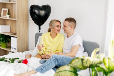 Genç bir adam ve bir kız yatak odasında bir kadeh şarap, bir balon ve çiçeklerle sevgililer gününü veya yıldönümünü kutluyorlar.