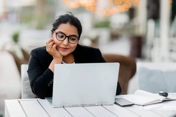 Uma jovem e confiante mulher indiana asiática sorri enquanto está sentada e trabalhando em seu laptop em um café elegante na rua em um dia de verão. — Fotografia de Stock