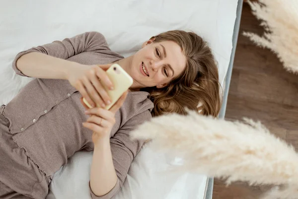 Uma jovem está deitada na cama em casa e navega pelas redes sociais em seu smartphone — Fotografia de Stock