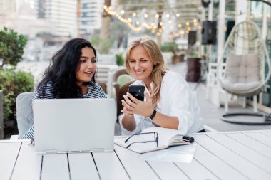 İki genç kadın, Avrupalı ve Hintli kızlar, bir kafe molasında gülüyorlar, telefonda selfie çekiyorlar, işe ara veriyorlar. İş, ofis dışında çalışmak, mola..