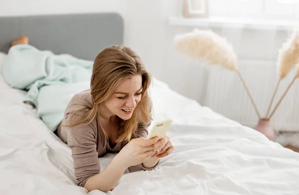 Jovem mulher alegre deitada na cama em casa e falando no smartphone — Fotografia de Stock