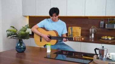 Gülümseyen Asyalı genç adam öğretmeniyle gitar çalıyor. Ev mutfağı, akıllı telefondan uzaktan öğrenme dersi. Nazik, arkadaş canlısı öğrenci. Yavaş çekim, boşluğu metin için kopyala