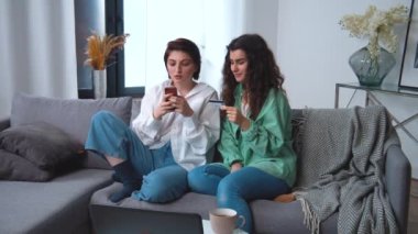 Lezbiyen çift internetten alışveriş yapıyor, banka kredi kartı ve akıllı telefonla ev oturma odasından ödeme yapıyor, gülümsüyor ve satın almaktan mutlu oluyorlar. Modern ödeme internet seçenekleri ve teknolojinin keyfini çıkarıyorum..