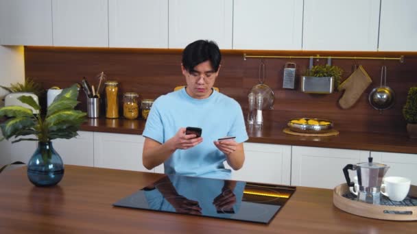 年轻的亚洲人坐在现代化厨房里 用智能手机用银行卡在网上购物 男人在网上购物后反应很快乐 在家里的公寓里中弹 — 图库视频影像