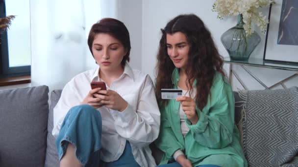 Лесбиянская пара делает покупки в Интернете, расплачивается банковской кредитной картой и смартфоном из гостиной дома, улыбается и с удовольствием покупает. Наслаждаясь современными возможностями и технологиями оплаты в Интернете — стоковое видео