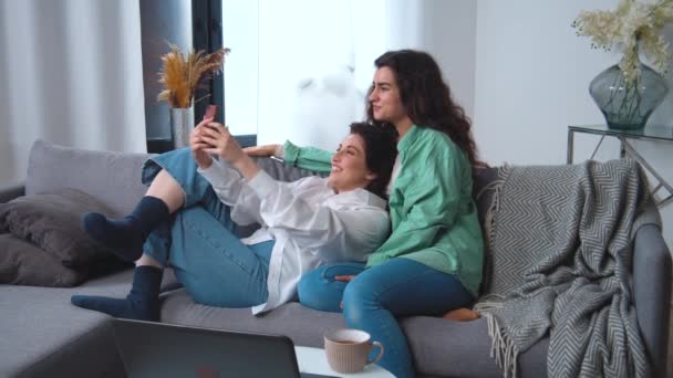 Happy lesbian couple is making a selfie or video call with a smart phone on couch in living room at home apartment. Receiving conference call — Stock Video © RomanMaidaniuk #535365404