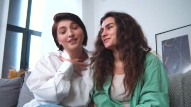 Lezbiyen çift görüntülü görüşme yapıyor, gülümsüyor, birbirlerine sarılıyorlar. İnsanlar çevrimiçi konuşmalar için akıllı teknolojiler kullanıyorlar. Oturma odasındaki kanepede oturmuş, duygusal bir şekilde gülüyorsun..