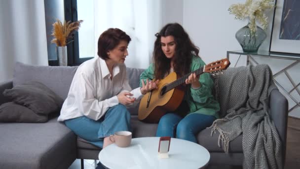 Лесбийская пара. Девочки учатся играть на гитаре, используя смартфон с онлайн учителем на диване. Дистанционные интернет технологии. Красивые улыбающиеся уверенные молодые кавказские этнические женщины красивое лицо. — стоковое видео