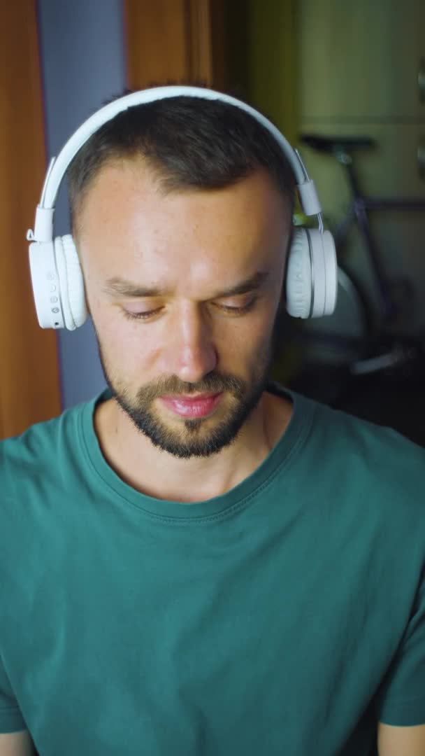 Retrato de jovem caucasiano bonito em fones de ouvido sem fio bluetooth ouvir música e olhando para a câmera. Um tipo sorridente em casa levanta a cabeça. Movimento lento vertical — Vídeo de Stock