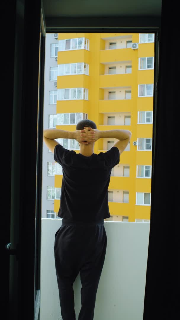 Jeune homme caucasien debout sur le balcon. mains sur la tête. Une tenue décontractée noire. Concept de solitude urbaine. Debout, dos à la caméra. — Video