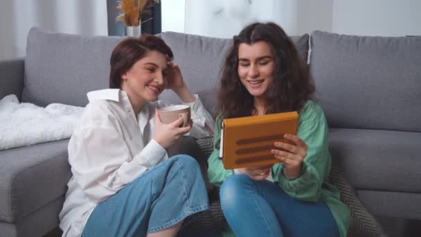 Happy young lesbian couple sitting on home floor, using tablet computer, browsing, laughing at funny photos, videos, having fun together, enjoying their weekend at home. Caucasian women. — Vídeo de Stock