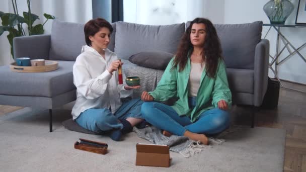 Расслабленная молодая лесбийская пара медитирует в позе лотоса, сидя на ковре с закрытыми глазами дома, наслаждаясь медитацией. Используя тибетский колокол. Онлайн медитация планшетный компьютер, психиатрическая помощь. — стоковое видео