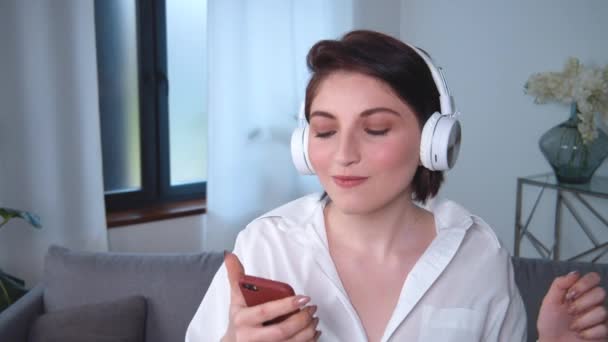 Портрет молодої білої жінки, що одягає бездротові навушники, слухає музику, вмикає пісню на смартфон. Веселі танці вдома. Красива дівчина насолоджується технологіями Bluetooth — стокове відео