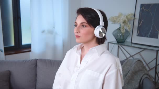 Portrait of young caucasian calm woman in wireless headphones listening to music or enjoying audiobook. Standing at apartment living room. Pretty girl enjoys bluetooth technologies — Wideo stockowe