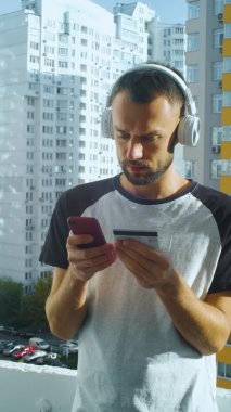 Bearded young man shopping online while standing at balcony, in wireless headphones. Vertical video of serious male person. Caucasian guy makes purchase and smiling, outdoor, urban city.