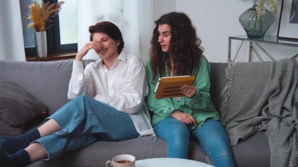 Glada unga lesbiska par sitter på soffan, använder surfplatta dator, bläddrar och skrattar åt roliga bilder, videor, ha kul tillsammans, njuta av sin helg hemma. Kaukasiska kvinnor skriker. — Stockvideo