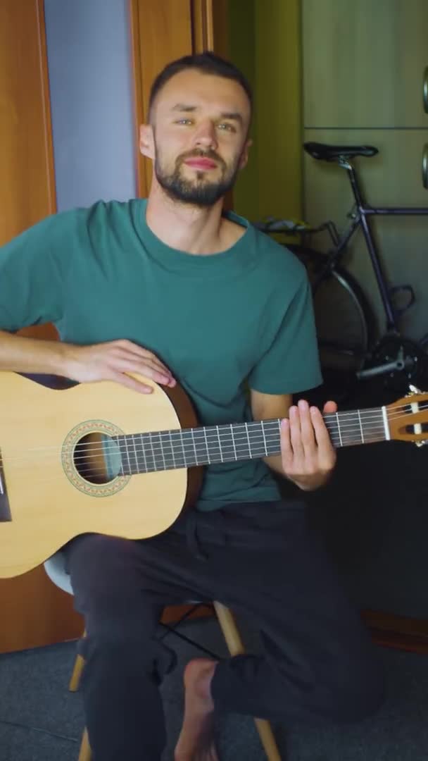 Retrato vertical del músico caucásico en casa. macho con guitarra sentado en el salón. estilo de vida músico europeo hombre en ropa casual mirando a la cámara — Vídeo de stock