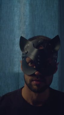 Kedi maskeli sakallı beyaz adam kameraya bakıyor ve ayağa kalkıyor. Bdsm seksi rol oyunu. Seks blogcusu ve koçun dikey görüntüleri. Düşük ışık, karanlık atmosfer.