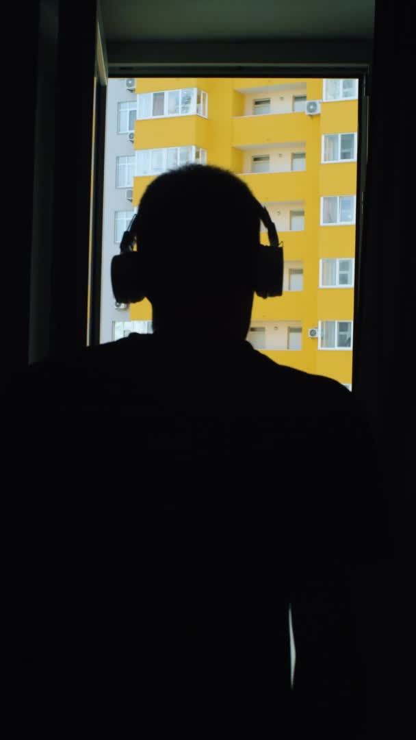 Vertiacal man goes through camera to balcony, looking at camera. male in wireless headphones listening music and posing for his blog, setting and checks video camera settings. — Stock Video