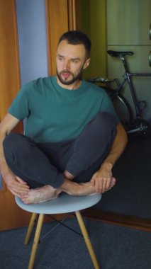 dikey modern hipster adam, modern koltukta lotus meditasyon pozisyonunda oturmaya çalışıyor. Kafkas erkek dinlenmek istiyor ve bacaklarında ağrı hissediyor..