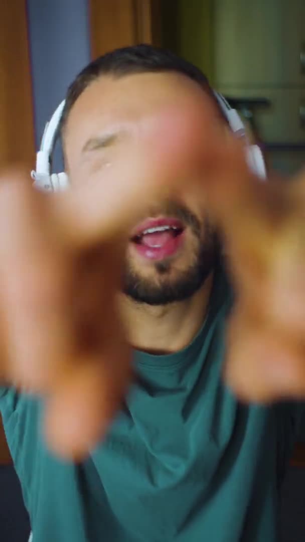 Portrait vertical homme barbu danse drôle dans les écouteurs sans fil et agitant les mains devant la caméra. blogueur gars regarde le public. caucasien blanc mâle obtenir plaisir de la musique et le chant. — Video