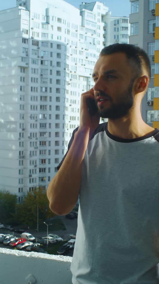 Uomo americano verticale riceve telefonate, chiacchiere allegre e sorridenti, in piedi sul balcone di casa. maschio barbuto caucasico gode di tecnologie a distanza wireless. giornata estiva soleggiata. — Video Stock