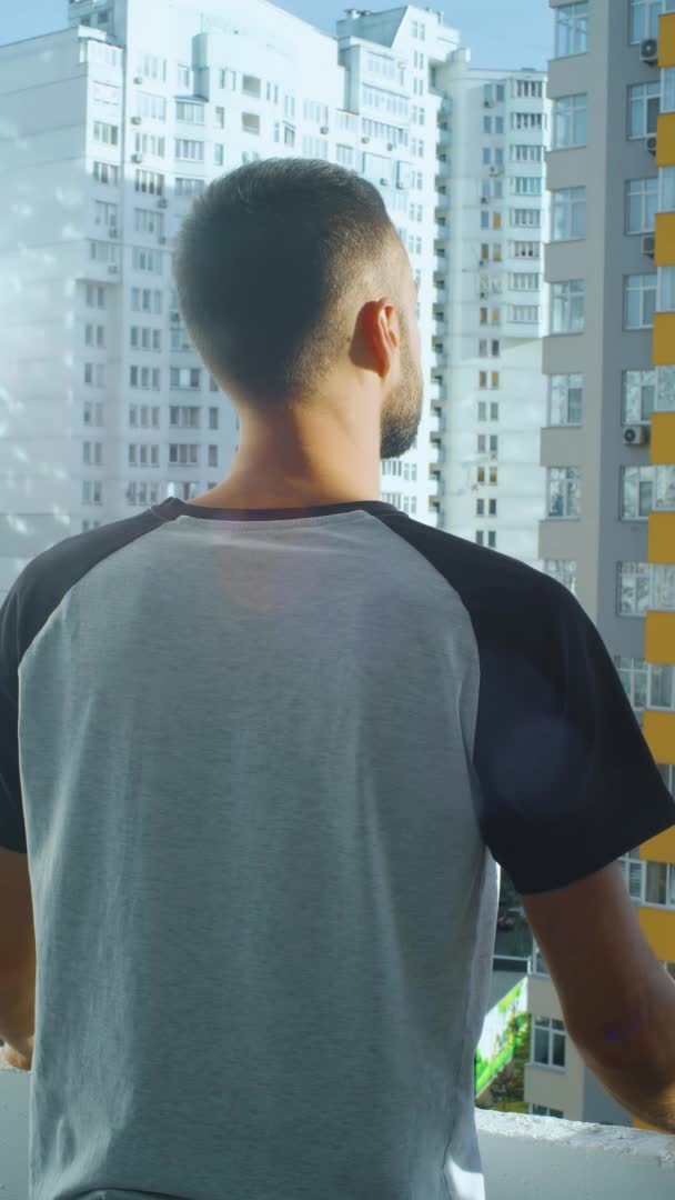 Vertical video of a man looking outside balcony, look at urban landscape. the caucasian man stands alone on the city balcony. young european male back view. sunny summer day outside. — Stock Video