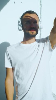 Dışarıda kablosuz kulaklık takan bir adamın dikey görüntüsü. Güneşe ellerle bakmaya çalışmak, gözlere gölge düşürmek. Sıcak yaz güneşli gününün atmosferi. beyaz sakallı beyaz erkek..