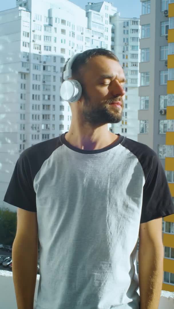 Bearded young man smiling on camera while standing on the balcony, and crossing arms. Vertical portrait of positive male person. Caucasian guy in wireless headphones outdoor, urban city — Stock Video