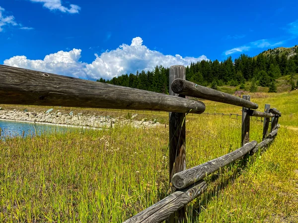 Beautiful view over a wooden fence between wooden fence posts, framed by a tall Alps Pine tree mountain forest backdrop. Country basic handmade rural wooden fence near lake, blue sky backdrop