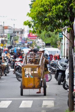 Yağmurdan sonraki bulutlu bir sabahta asfalt yolda arabasını çeken çalışkan bir çöpçü. Solo - Endonezya. 9 Aralık 2021