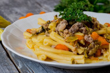 Kabaklı makarna, kabak çiçekleri, sarı domates ve beyaz tabakta servis edilen sosis.