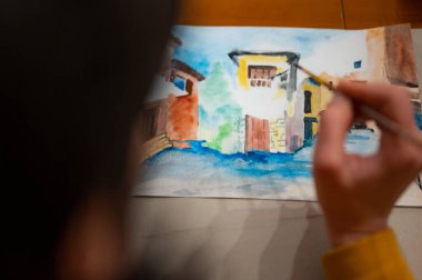 Yaratıcı sanat atölyesinin işleyişi. Ressam, fırça ve suluboya kullanarak kağıt üzerine renkli mimari şehir manzarası çiziyor