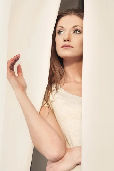 Beautiful girl looking through the curtains — Stock Photo, Image