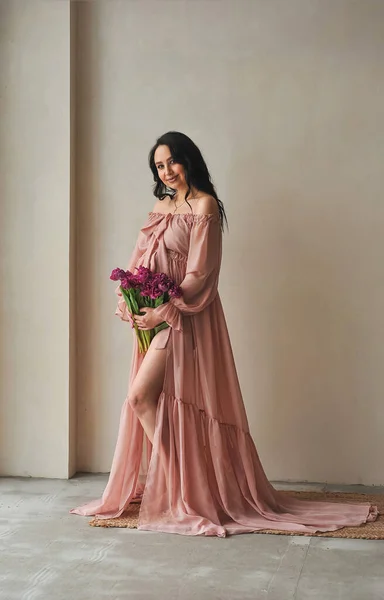 Güzel esmer, pembe elbiseli hamile kadın elinde bir buket menekşe laleyle gülümsüyor. Hamilelik fotosferi