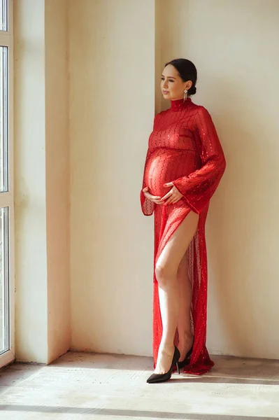 在摄影棚里 穿着漂亮闪亮的红色连衣裙的漂亮孕妇站在墙边 抱着怀孕的肚子 望着窗外的画像 妊娠期光程 — 图库照片