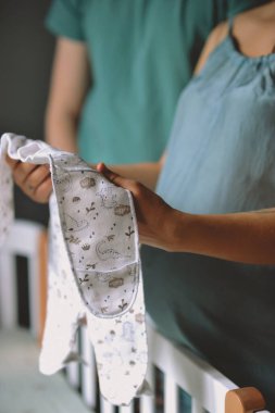 Mavi pijamalı hamile bir kadın beşiğin yanında duruyor ve elinde bebek kıyafeti var. Aile kavramı. Anne babalar bebek bekliyor.