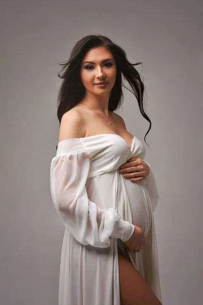 Beyaz elbiseli, hamile karnını tutan, siklonamada izole edilmiş çekici hamile kadın portresi. Bebek bekliyormuş. Saçların dalgalanır.