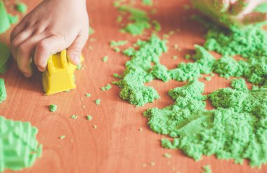 Kahverengi masanın üzerinde yeşil kinetik kumla oynayan çocuğun ellerini kapat; seçici odak