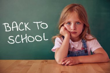 School child (girl) in classroom. Funny sad kid against green chalkboard. Idea and creativity concept. Copyspace on background. Back to school concept. 